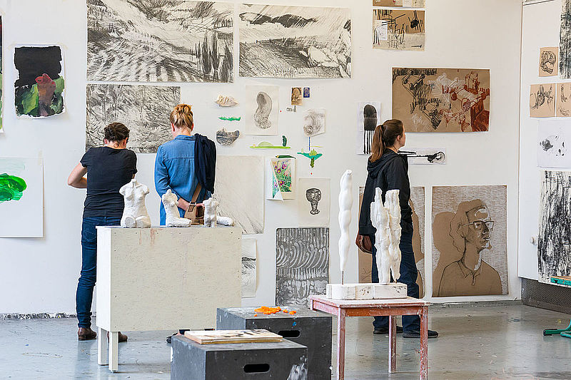 Drei Personen sehen sich die Arbeiten von Studierenden in der Jahresausstellung an.